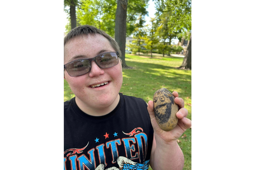 A camper smiling at the camera while holding up a rock with a face drawn on it