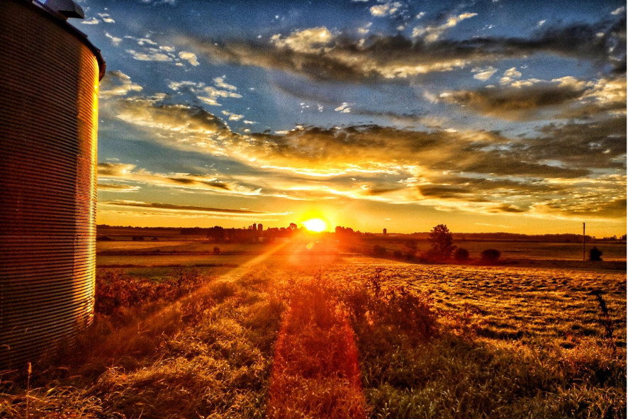 A sun setting on the horizon of a farm field with a silo on the lefthand side of the frame