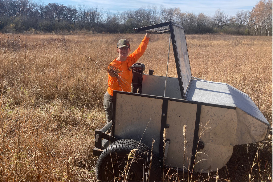 A person standing behind a pull-behind seed stripper for collecting local seeds to plant and to interseed lower diversity prairie and old fields.