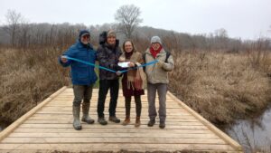 NRF board members standing with staff from The Prairie Enthusiasts on a newly built bridge above a stream, holding a ribbon and a check. 