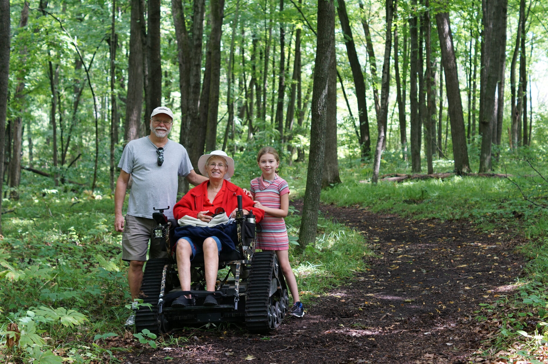 person sitting in an all-terrain wheelchair and two people standing behind them on a trail in a forest